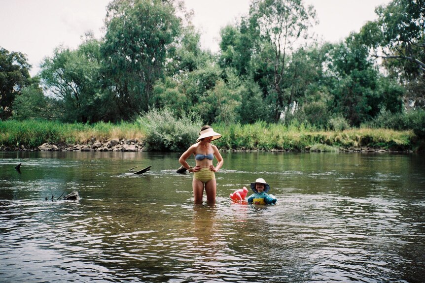 Singer Fanny Lumsden swimming in a river in regional NSW, accompanying her child in floaties. Both wearing sun hats.