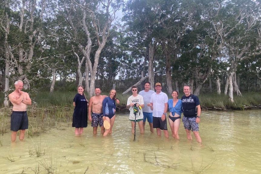 A group of people stand in shallow river water, with trees and reeds behind them.