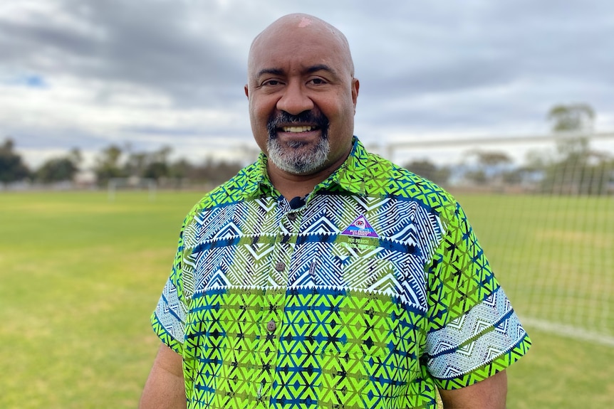 A smiling,  bald Pacific Islander man with a grey beard wearing a green and blue heavily patterned shirt.