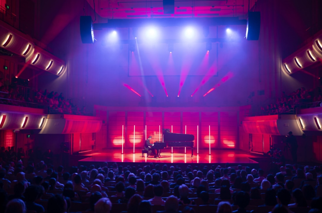 CJ Jones plays a grand piano on a brightly lit stage in a modern concert hall before a large seated audience