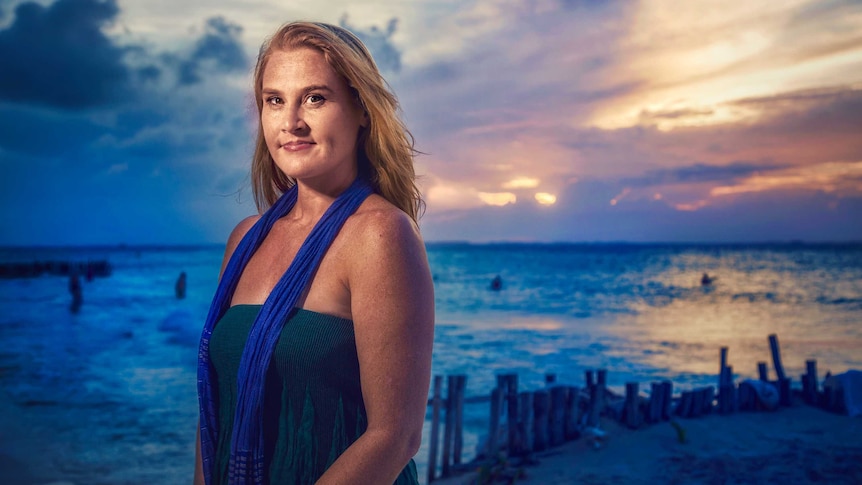 Wendy Dent looking at the camera wearing a blue sleeveless dress standing on beach with the sun shining behind the clouds