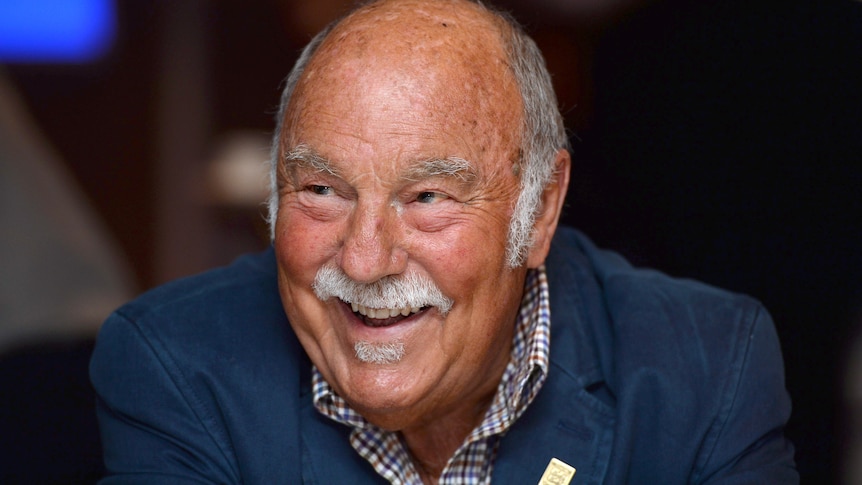 An elderly man with moustache and receding hair wearing dark suit smiles in a room.