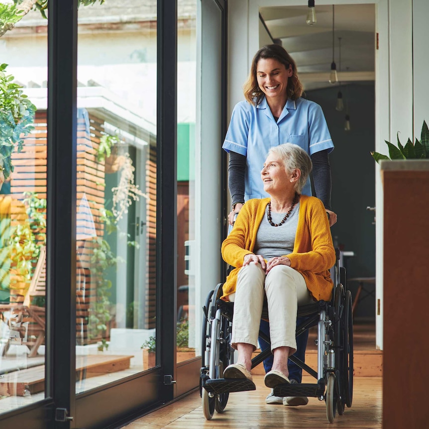 An elderly woman smiles looking out the window, while being moved in a wheelchair by a young nurse