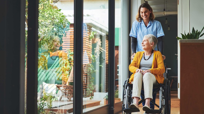 An elderly woman smiles looking out the window, while being moved in a wheelchair by a young nurse