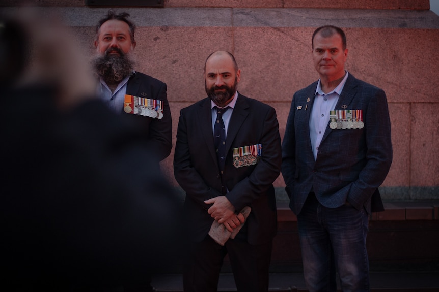 Three former service men with medals on their jackets stand in front of the Cenotaph.