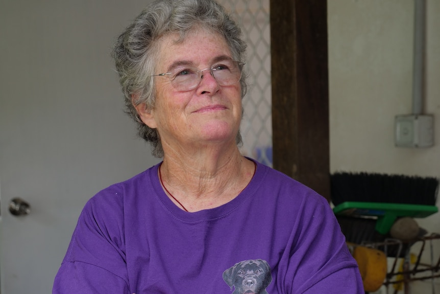 Grey-haired woman wearing purple shirt, wearing glasses in front of white cabin.