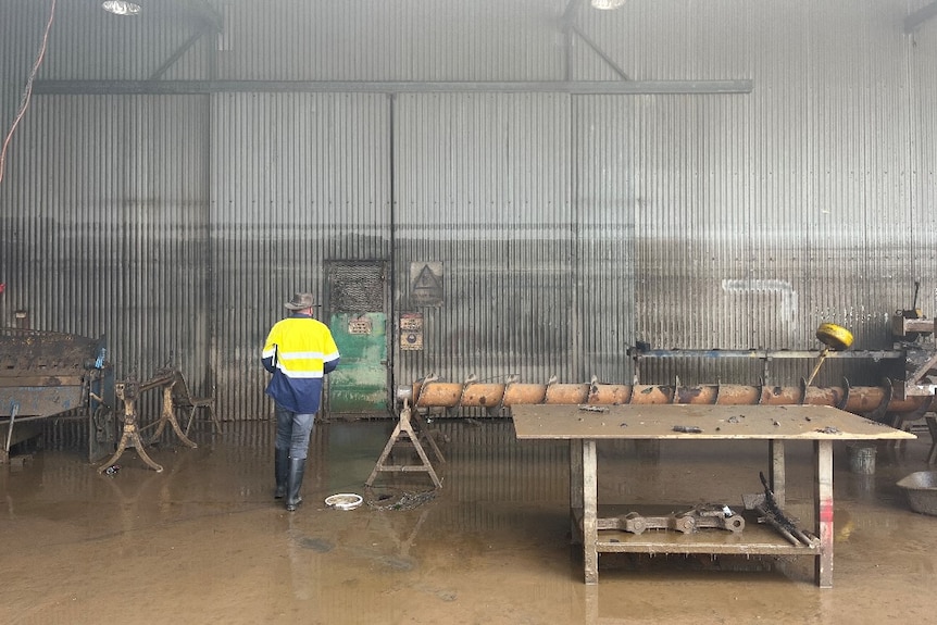 A worker in high-viz jacket walks through a shallow flooded shed with a table and other equipment.