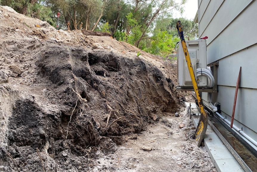 A trench has been dug on the border of a home roughly 60 cm high. A shovel is standing stuck in the dirt. 