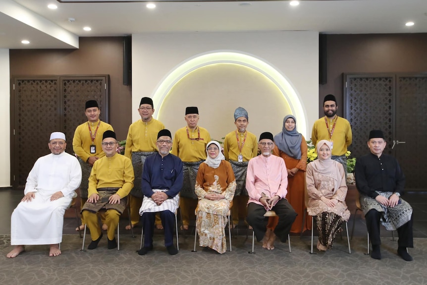 A group of 15 people in two rows in traditonal Muslim garb pose for photo in decorative room. 