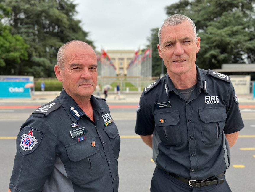 Two men in navy fire fighter uniforms stand outside the United Nations row of flags 