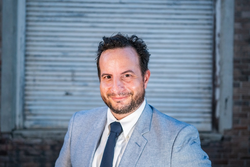 Comedian Dan Ilic from A Rational Fear wearing a grey suit and smiling.