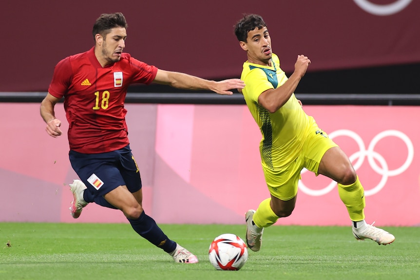 A man wearing yellow runs past a man wearing red towards a soccer ball