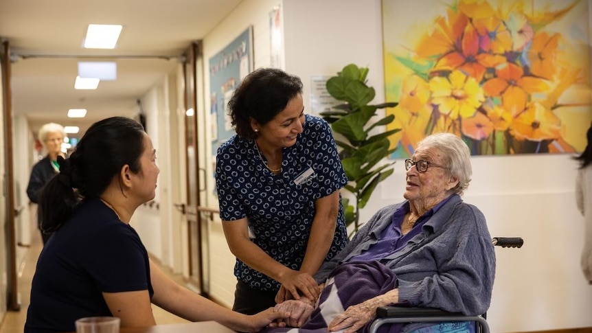 An elderly woman in a wheelchair speaks to two smiling female staff in an aged care facility
