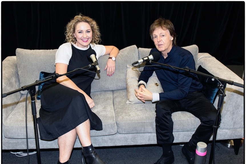Rowe and McCartney sitting on a couch with microphones on stands in front of them.