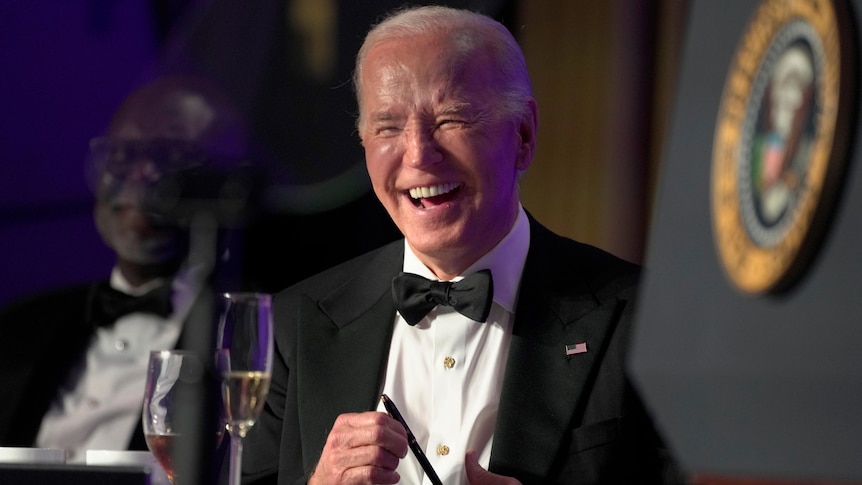 Joe Biden sits, laughing and hokding a pen, with a glass of sparkling wine in front of him. He's in black tie. 