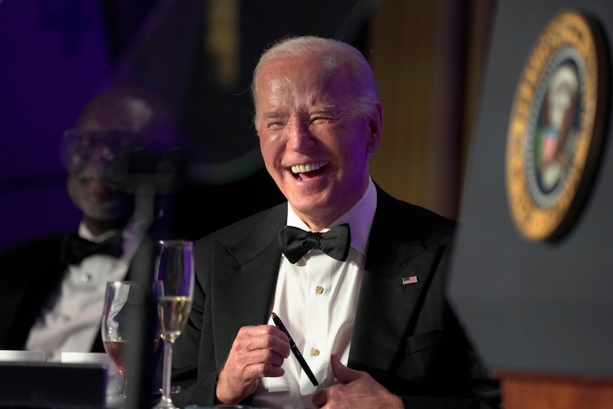 Joe Biden sits, laughing and hokding a pen, with a glass of sparkling wine in front of him. He's in black tie. 