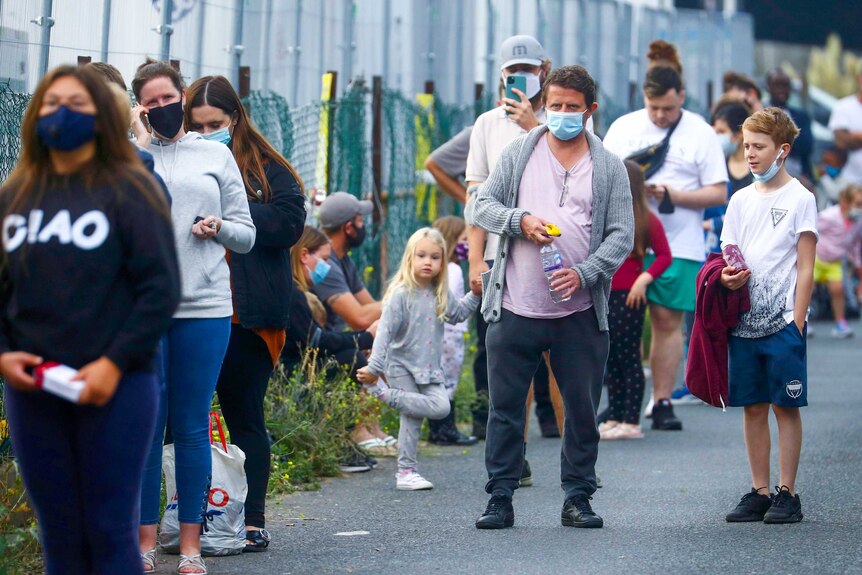 A line of people along a fence, some wearing face masks