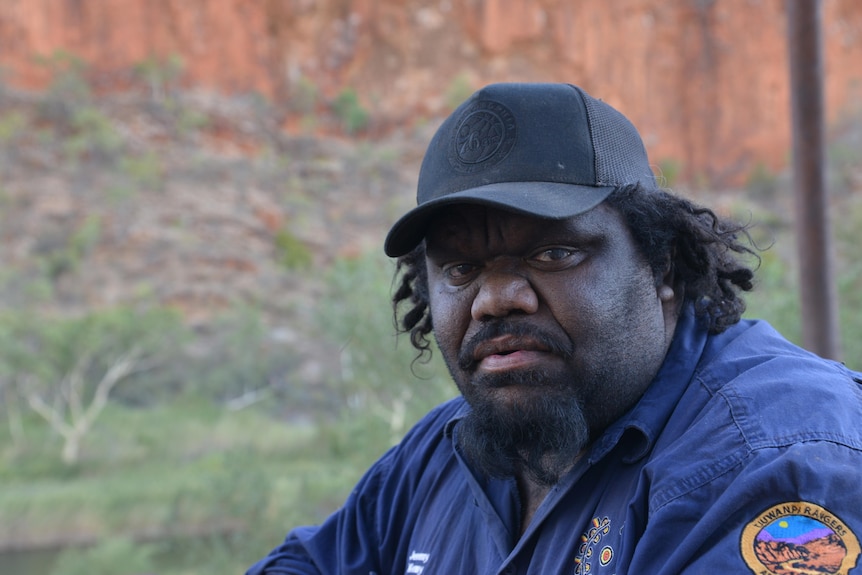 A man in a blue collared work shirt and black cap looks directly at the camera.