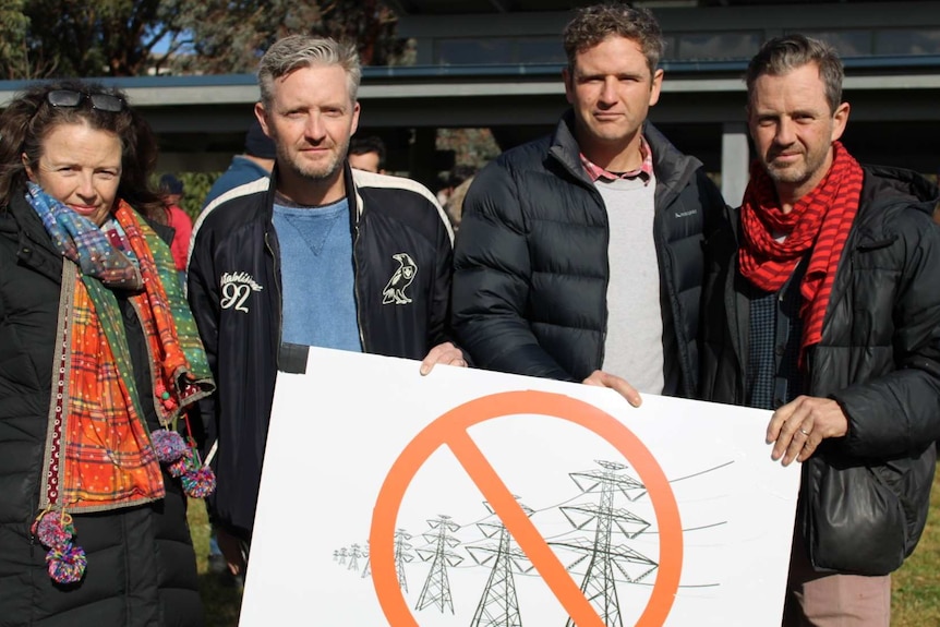 Four people standing with a sign protesting against mega powerlines at a community meeting