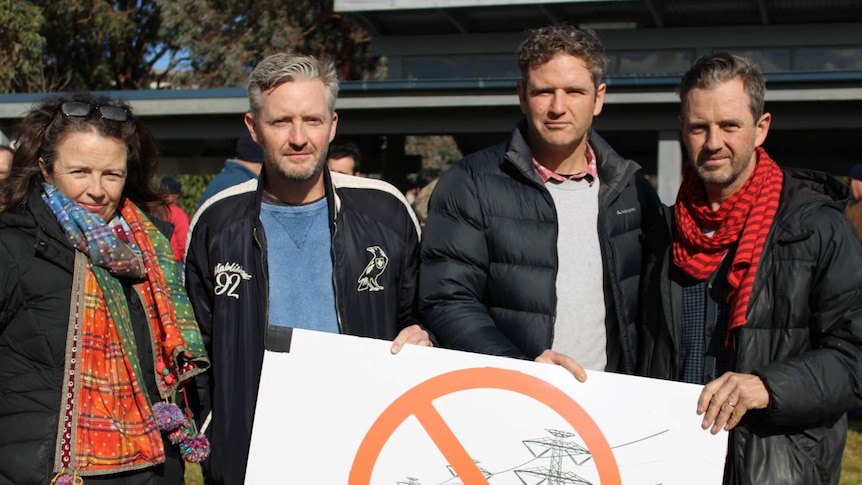 Four people standing with a sign protesting against mega powerlines at a community meeting