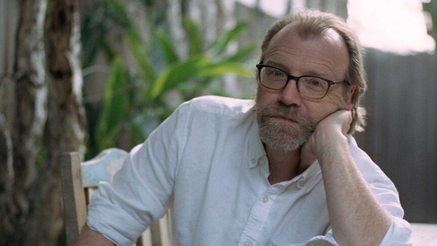 Author photo of George Saunders wearing button up white shirt, sitting on chair at desk outside.