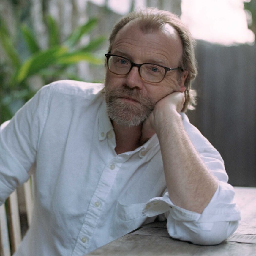 Author photo of George Saunders wearing button up white shirt, sitting on chair at desk outside.