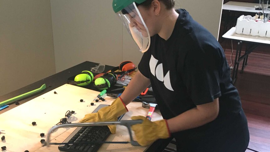 A woman wearing safety equipment uses a bowsaw on a computer keyboard.