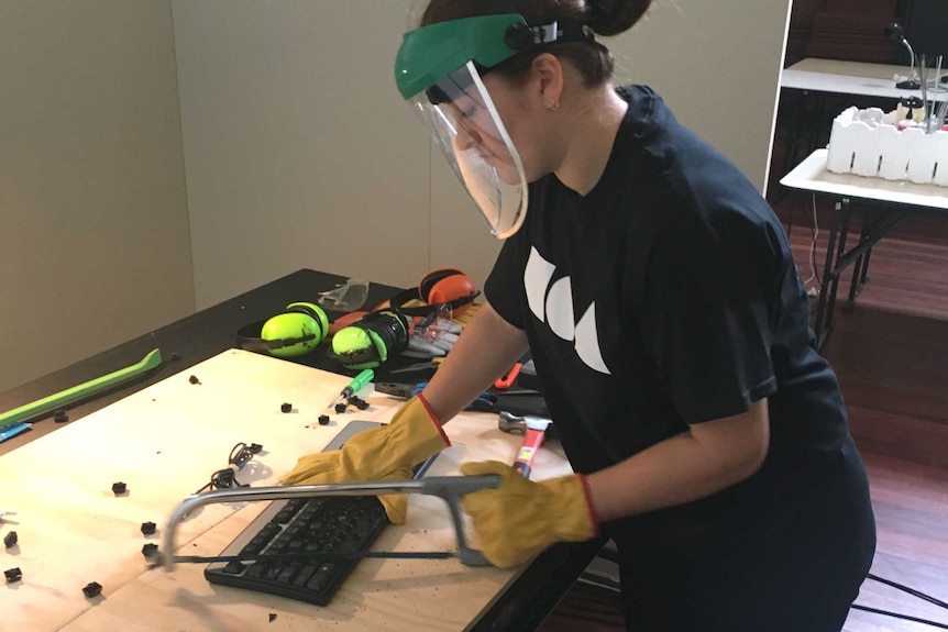 A woman wearing safety equipment uses a bowsaw on a computer keyboard.