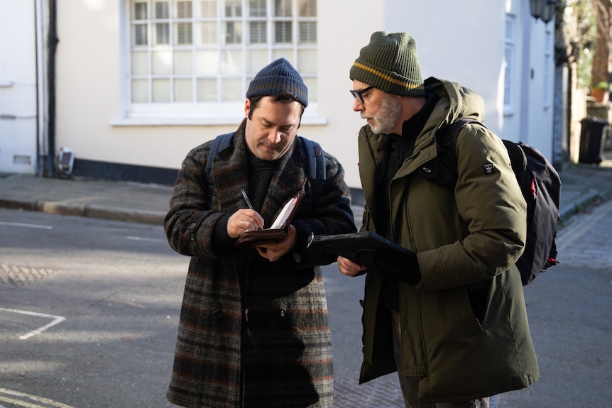 A man in a beanie and overcoat writes notes in a book while talking to a similarly dressed man.