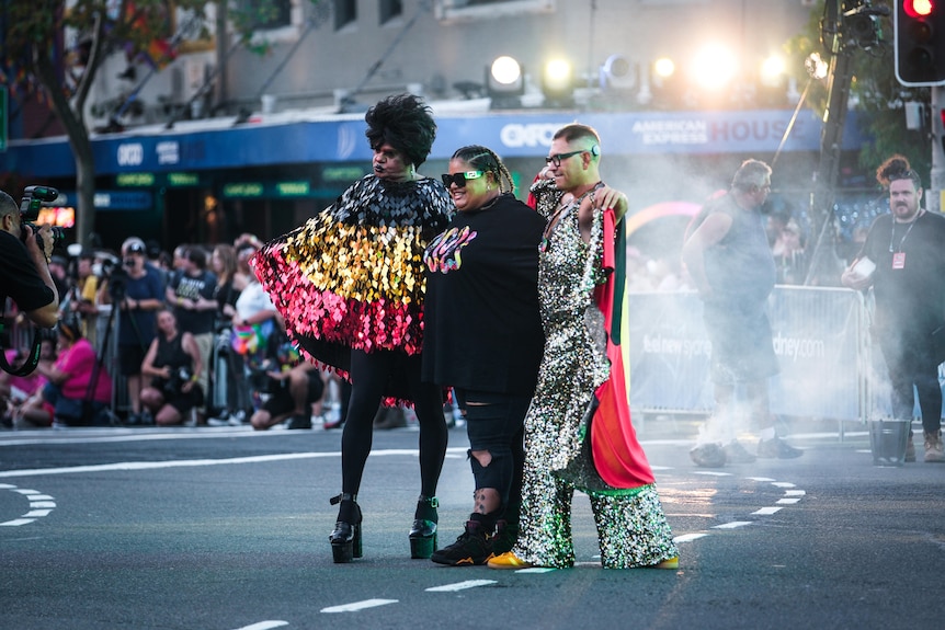 Trois personnes vêtues de vêtements scintillants aux couleurs du drapeau aborigène posent dans une rue