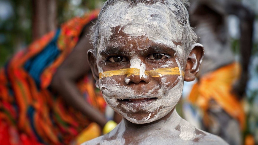 A young aboriginal boy painted in white and yellow body paint stares into the camera.