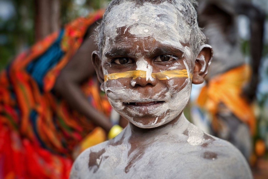A young aboriginal boy painted in white and yellow body paint stares into the camera.