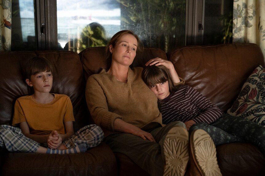White middle-aged mother wearing brown shirt sits between two pre-teen sons on a couch in a living room watching a TV.