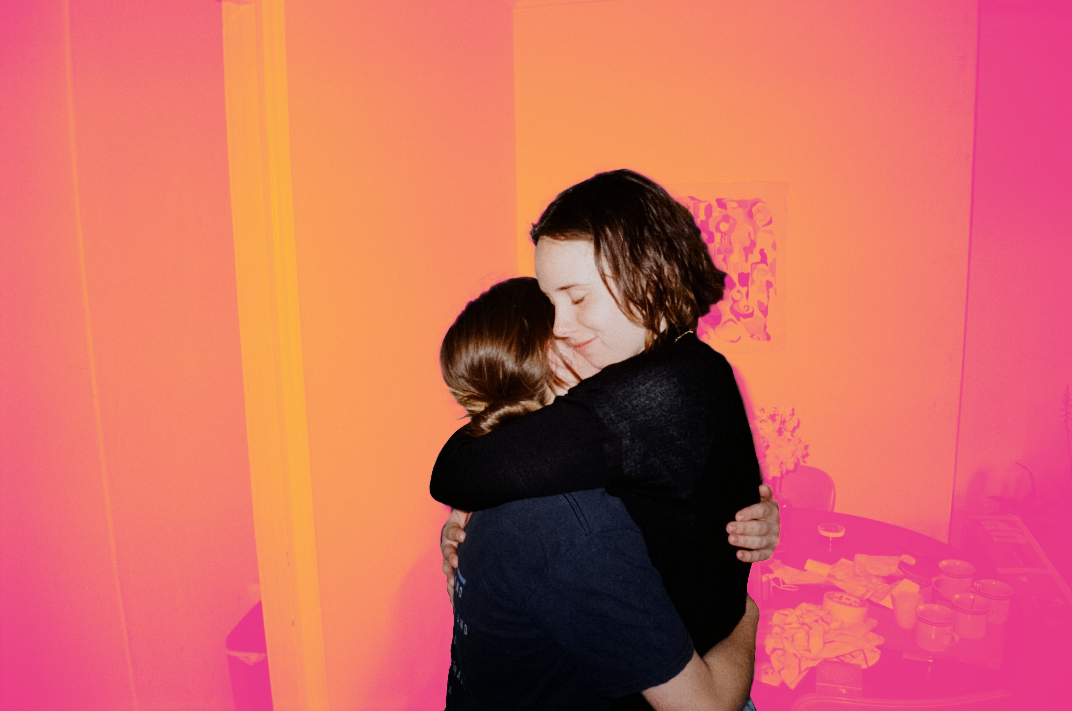 Two young adults hug very tightly, their eyes closed, the background has been manipulated to be a hazy orange and pink