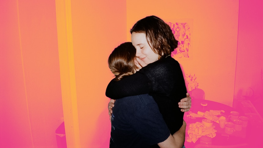 Two young adults hug very tightly, their eyes closed, the background has been manipulated to be a hazy orange and pink