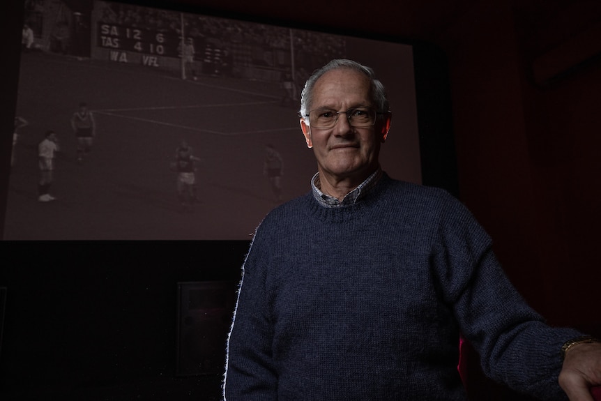 A man looks at the camera, an old football game projected on the screen behind him.