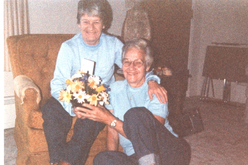 Two women seat embracing, one on an armchair, the other on the floor next to her, both smiling widely.