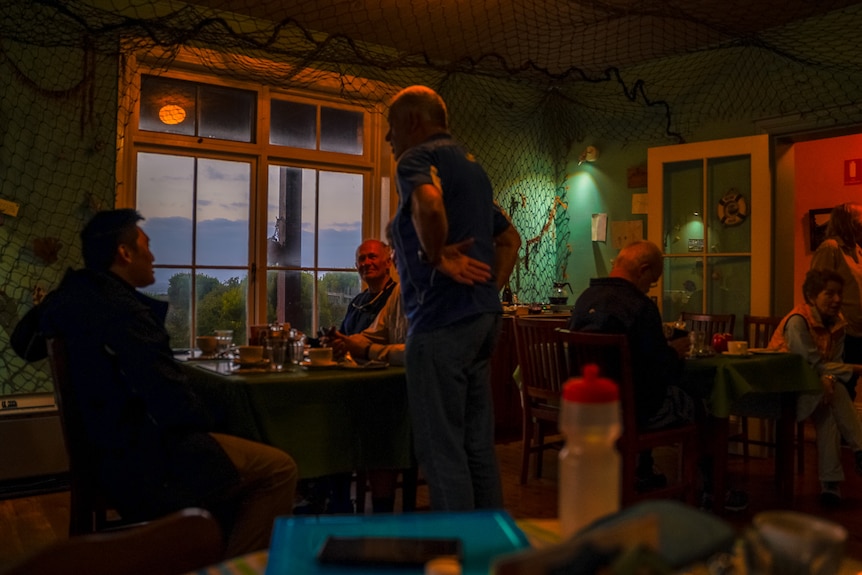 Three men sitting at a table in a dimly lit dining area talk to each other.