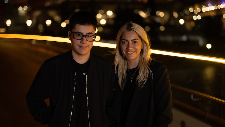 A teenage boy with glasses stands next to a woman, pictured at night with lights behind them