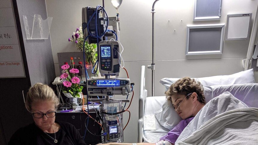 Ned Desbrow asleep in a hospital bed and his mother Melita Carlyon sits in a chair nearby.