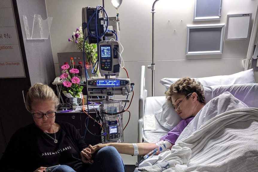 Ned Desbrow asleep in a hospital bed and his mother Melita Carlyon sits in a chair nearby.