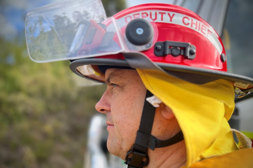 Male firefighter looks straight ahead wearing red helmet and yellow protective uniform. Picture taken from side angle. 