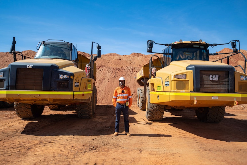 A man stands between two giant mining trucks.