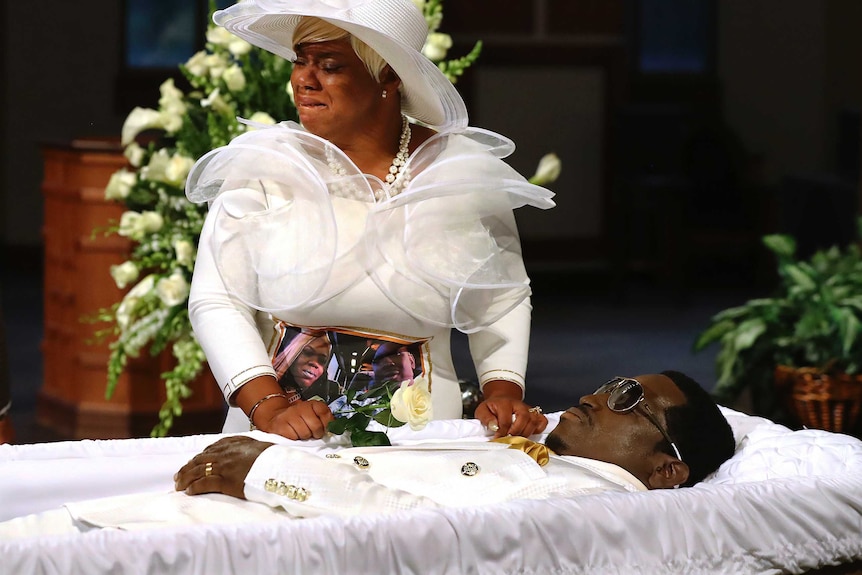 A woman wearing all white and a white hat cried over a man dressed in all white, who is laid in an open casket.