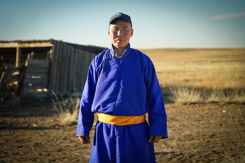 A man wearing a blue robe with yellow belt around the middle, and a cap on his head, stands in front of a shed