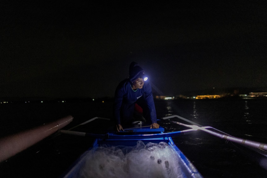 A man wears a torch in the dark and leans on top of a small boat in the water. 