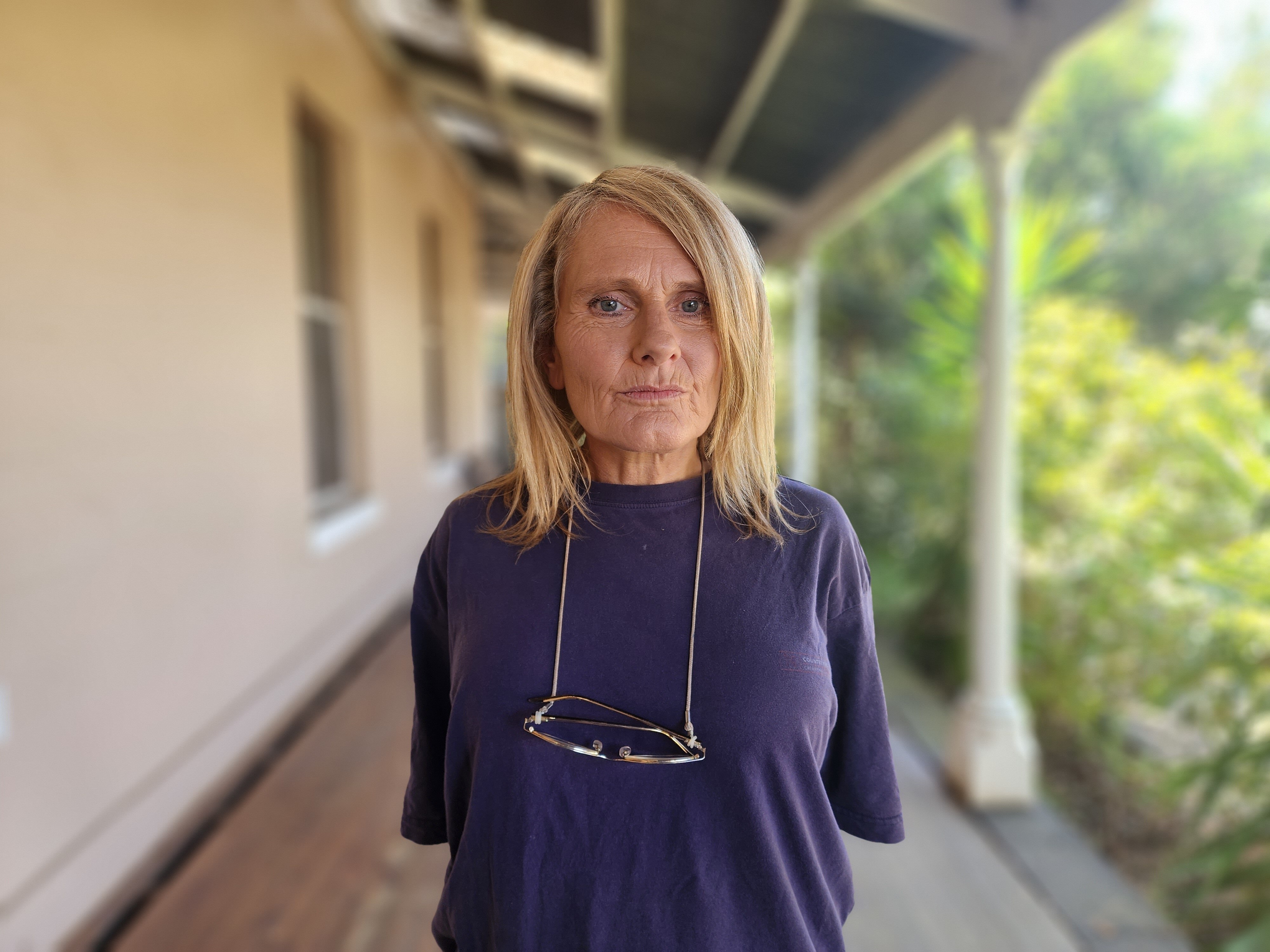 A lady standing in a blue t-shirt with reading glasses hanging around her neck 