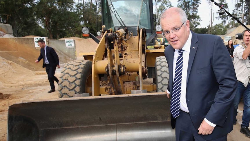 Scott Morrison poses in front of a digger while a camera operator positions a boom mic above him and a man in a suit walks past.