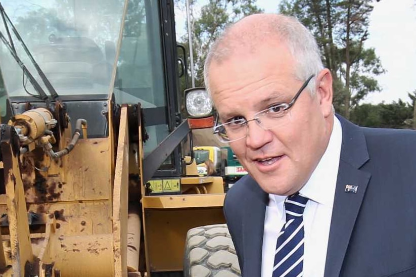 Scott Morrison poses in front of a digger while a camera operator positions a boom mic above him and a man in a suit walks past.