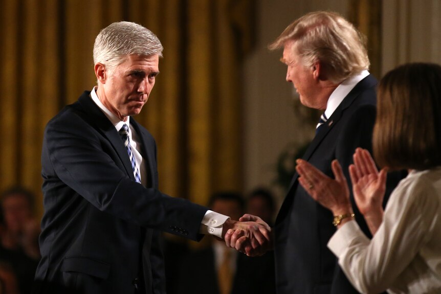 A middle-aged man with grey hair in a suit shakes hands with Donald Trump as a woman applauds.